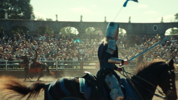 A screenshot from the series Ren Faire. Two knights on horseback, one in the foreground and other in the distance, duel in front of a crowd seated on bleachers.